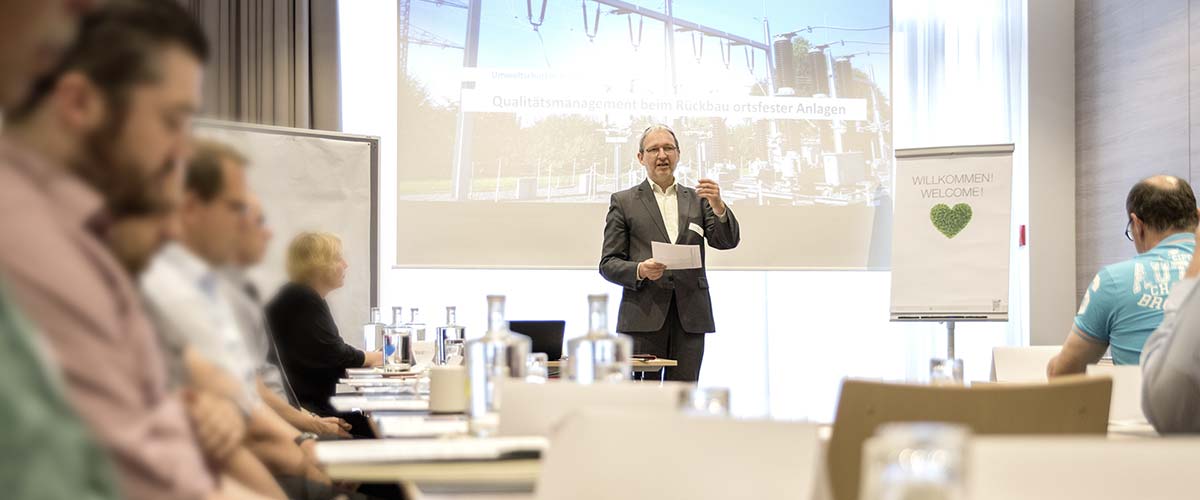 Sachverständiger Dr. Schoeps steht vor einer großen Beamerleinwand und hält einen Vortrag vor an mehreren Tischen sitzenden Kunden